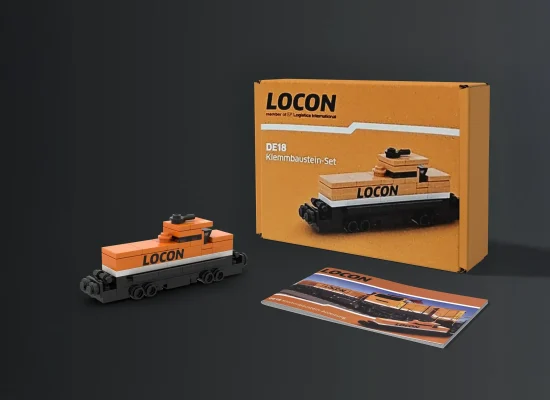 Eine kleine orange-schwarze Spielzeugeisenbahn aus Klemmbausteinen mit der Aufschrift LOCON steht neben der dazugehörigen Schachtel und einem Anleitungsheft vor einem dunklen Hintergrund.